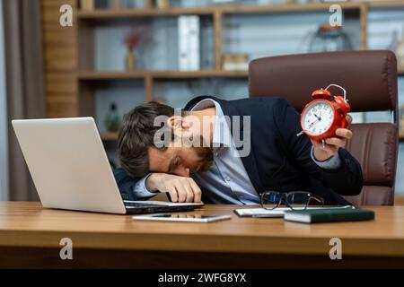 Tired office manager laying down on folded hand on table with personal gadgets and holding red alarm clock. Overworked man having insomnia or chronic fatigue syndrome and taking nap during busy day. Stock Photo