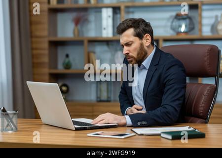 Unhealthy businessperson holding hand on stomach and grimacing in pain while sitting in workspace interior. Handsome male feeling unwell and having severe belly ache due to skipping full meals. Stock Photo