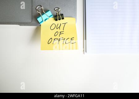 Message Out Of Office stick on top of computer laptop next to notepad on office desk. Top view. Stock Photo