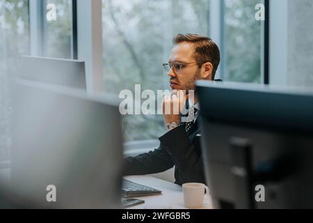 Young businessman with hand on chin using computer at desk in corporate office Stock Photo