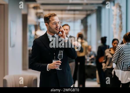 Young businessman holding champagne flute while talking on mobile phone during business conference Stock Photo