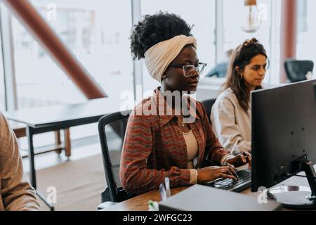 Young businesswoman wearing headscarf and eyeglasses using computer while sitting by female colleague at desk in office Stock Photo