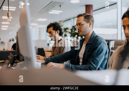 Young businessman using computer while working amidst colleagues sitting at desk in office Stock Photo