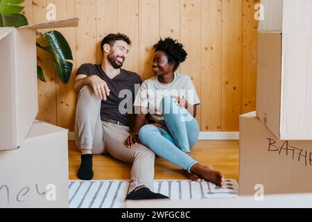 Happy multiracial couple talking while sitting together near wooden wall at new home Stock Photo