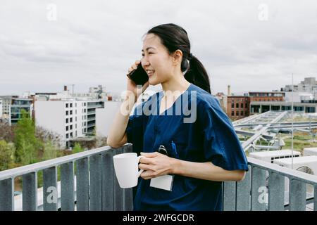 Smiling mid adult female nurse holding coffee cup while talking on smart phone Stock Photo