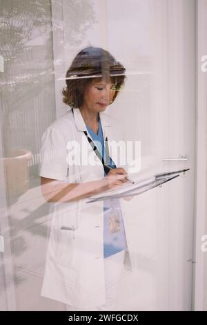 Senior female physician examining medical record seen through glass Stock Photo