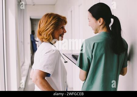 Happy female healthcare workers looking at each other while standing in hospital corridor Stock Photo