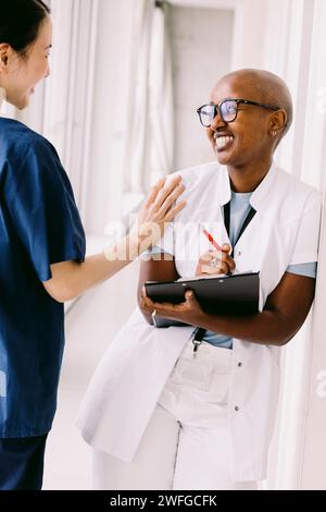Happy female doctor and leaning with clipboard while looking at colleague in hospital corridor Stock Photo