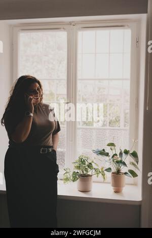 Smiling businesswoman talking on smart phone while standing near window at office Stock Photo