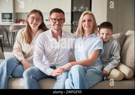 Big family chilling together on cream sofa. Cute family portrait of four members looking to the camera. Happy moments of close people in modern living room. Stock Photo