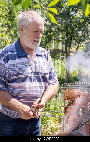 A gray-haired man is grilling meat on a grill by the brazier. Meat is cooked on a homemade barbecue in the nature. Stock Photo