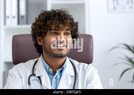 A young male Hindu doctor with curly hair smiling in a medical office, representing professionalism and care. Stock Photo