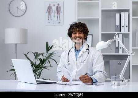 Confident young medical professional with stethoscope seated at a desk, using laptop in a bright clinic office. Stock Photo