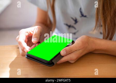 Young woman in blue home tee shirt using her smartphone with green screen on table, scrolling news, photos or communicating in social networks. Online Stock Photo