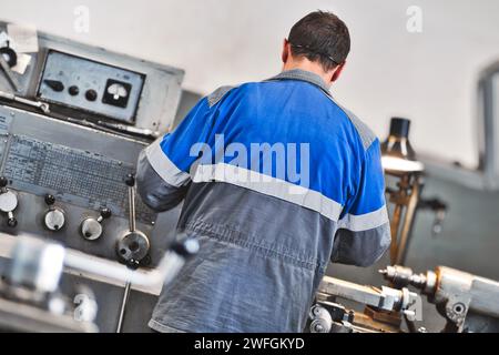 Turner in overalls works at lathe in turning shop at factory. Elderly worker turns metal part on machine. Rear view from back. Stock Photo