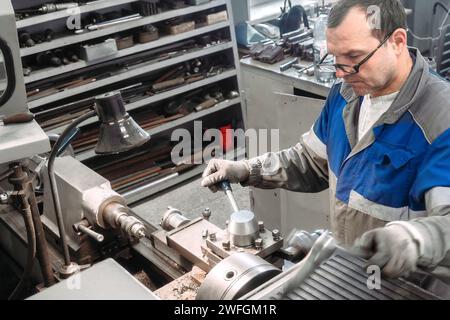 Portrait of professional turner at work on lathe in workshop. 50-55 year old turner in overalls and glasses turns part in workshop on machine. Photography authentic work process. Stock Photo