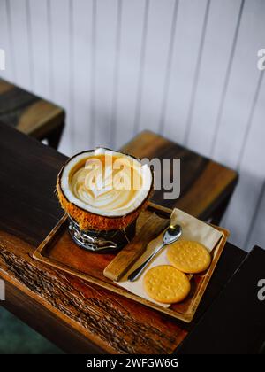 Hot Latte coffee in coconut. Tray with cup of tasty coconut coffee. Cup of tasty coconut coffee and spoon with Crackers on wooden table. Warm cafe lat Stock Photo
