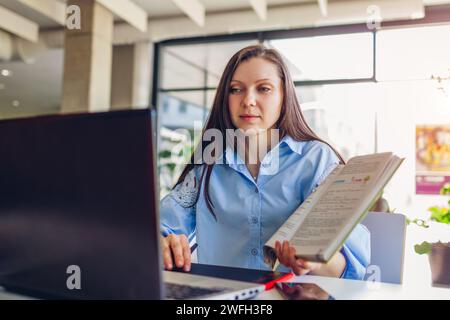 Young female teacher working through videoconference in office using laptop. Woman showing students book online. Remote lesson Stock Photo