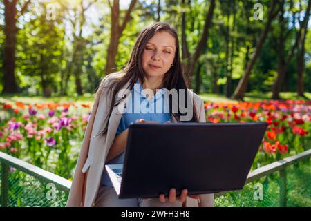 Outdoor portrait of young business woman working in spring park on laptop. Female entrepreneur uses internet in public garden Stock Photo