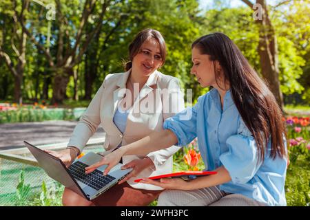 Two young women cowokers chatting in spring park discussing work. Teachers use notebooks to share ideas outdoors. Teamwork Stock Photo