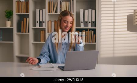 Multitasking Caucasian woman working with laptop writing in notebook talking mobile phone call loudspeaker happy businesswoman girl write notes typing Stock Photo