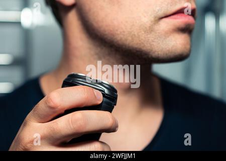 Shaving beard with machine. Man with electric shaver. Trimmer for face hair. Male grooming razor. Mirror in bathroom. Short stubble. Closeup of neck. Stock Photo