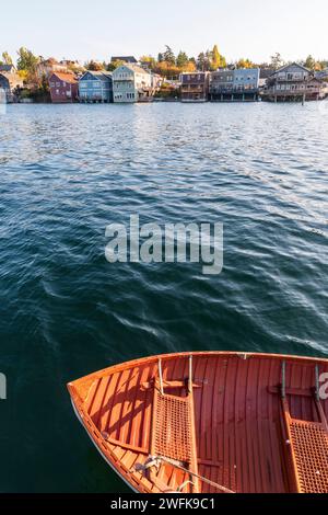 A vertical photo of a red-orange rowboat in the waters of Penn Cove with colorful buildings built on pilings along the waterfront of Coupeville, Whidb Stock Photo