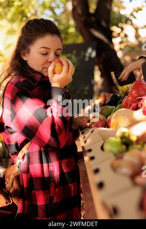 Female buyer enjoying natural fresh smell of apples, standing in front of farmers market stand. Woman smelling organic colorful fruits before buying homegrown eco produce from counter. Stock Photo