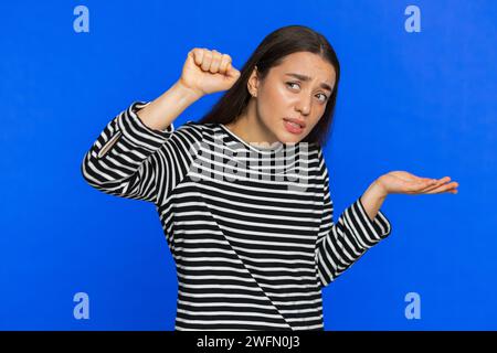 Knock-knock who is there. Confused excited Caucasian woman knocking door gesture asking who is at home feeling embarrassed, no idea being clueless and uncertain. Young girl isolated on blue background Stock Photo