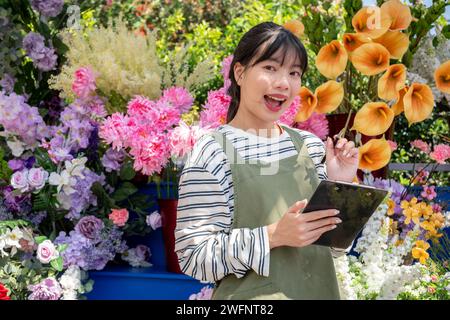 An attractive and charming young Asian female florist, or a small flower shop owner with a clipboard, is standing in front of her flower shop. Stock Photo