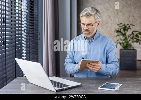 Thoughtful mature male with grey beard looking at portable computer screen while taking notes in domestic workplace. Attentive employee watching training webinar and making summary with main theses. Stock Photo