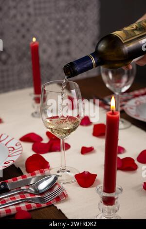 Hand serving a glass of white wine, on a dinner table set for two with red roses' petals. There is a lighted red candle. Vertical image Stock Photo