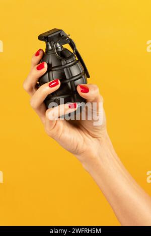 Hand of anonymous slim young female with manicured and nail polished fingers holding black metal grenade with safety pin on body filler with pull ring Stock Photo