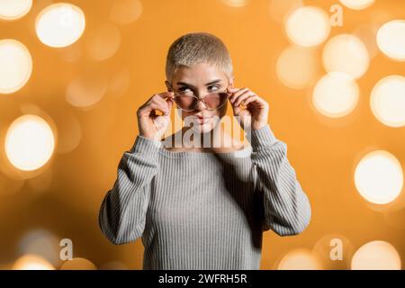 Young female blonde with shorthair looking away at corner while standing in full sleeves pullover with shade in light and adjusting sunglasses on nose Stock Photo