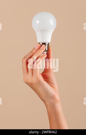 Hand of anonymous young female showing white LED light bulb in ecology concept against beige background in studio Stock Photo