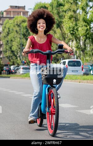 Full body of cheerful Moroccan woman riding electric bicycle on city street and looking forward with toothy smile Stock Photo
