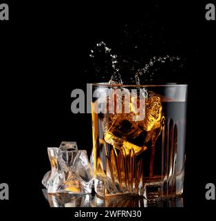 Glass with whiskey and falling ice cube with splashes on black background. Stock Photo