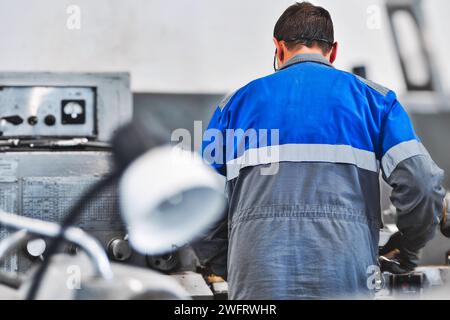 Portrait of professional turner at work on lathe in workshop. 50-55 year old turner in overalls and glasses turns part in workshop on machine. Photography authentic work process. Stock Photo