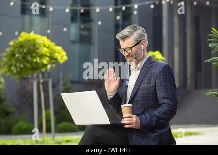 Experienced senior businessman in suit using laptop and greeting someone, standing outside a modern office building. Stock Photo