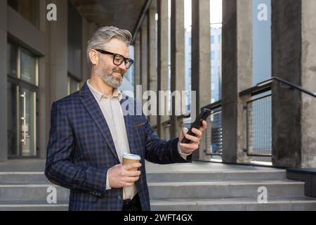 Cheerful mature businessman in stylish suit using smartphone and holding coffee outside an office building on a bright day. Stock Photo