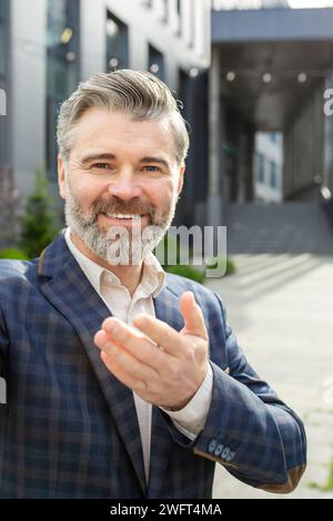 Cheerful experienced senior professional offering a handshake in a friendly gesture, standing confidently outside an office. Stock Photo