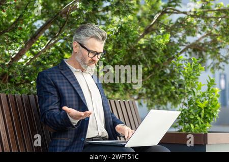 Mature male financier in a smart suit using a laptop outside an office building, exuding professionalism and experience. Stock Photo