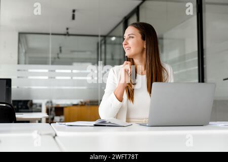 A businesswoman ponders with a smile, looking away from the laptop, contemplating a new idea, or reflecting on a successful deal. Concept of creative and strategic aspects of business thinking Stock Photo