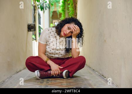 Front view of young Venezuelan Latina woman with curlers, sitting cross-legged on hallway floor, worried looking at camera Stock Photo