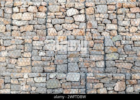 Stonewall behind wire mesh background texture. Metal lattice for protection in front of empty grey rocky wall construction backdrop. Copy space Stock Photo