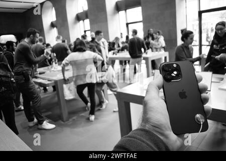 Paris, France - Sep 22, 2023: A man holds the latest Apple iPhone 14 Pro inside an Apple store, surrounded by curious customers who are chatting and coming to see the new smartphones. The image is in black and white Stock Photo