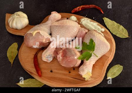 On a dark background, on a board, lie raw chicken legs ready for cooking. Next to the meat are bay leaves, pepper, and garlic. Stock Photo