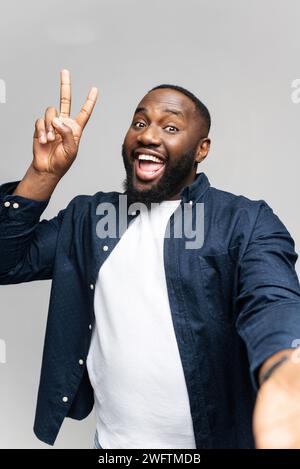 A cheerful African-American man is taking a selfie, flashing the peace sign with his fingers. His joyful expression and casual style convey a sense of fun and spontaneity. Stock Photo