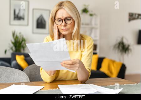 Concentrated upset senior woman sitting at the desk and looking at the documents deep in thoughts and doubt, disappointed and worried female freelancer has trouble and difficulties Stock Photo