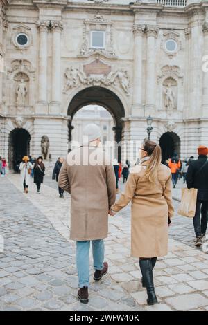 Two individuals stroll hand in hand amidst urban scenery, passing by a majestic structure Stock Photo